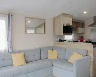 A cozy, modern interior of a mobile home featuring a gray sectional sofa with yellow cushions, a wooden kitchenette with a mirror, and a bright window.