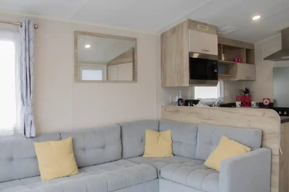 A cozy, modern interior of a mobile home featuring a gray sectional sofa with yellow cushions, a wooden kitchenette with a mirror, and a bright window.
