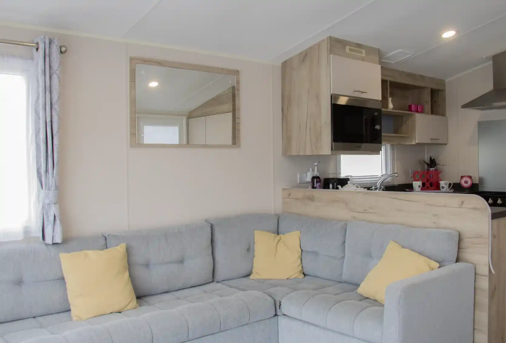 A cozy, modern interior of a mobile home featuring a gray sectional sofa with yellow cushions, a wooden kitchenette with a mirror, and a bright window.