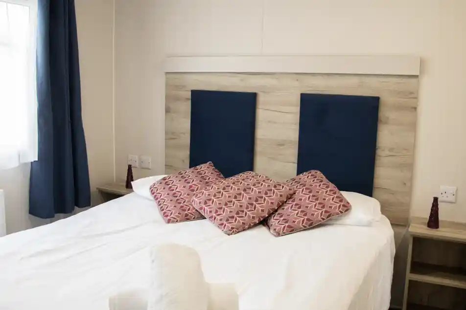 A neatly made bed with white linens, topped with decorative pillows featuring a geometric pattern. The headboard has a wood panel design with blue fabric sections. Two bedside tables with small red decorative items are visible, and curtains frame a window in the background.