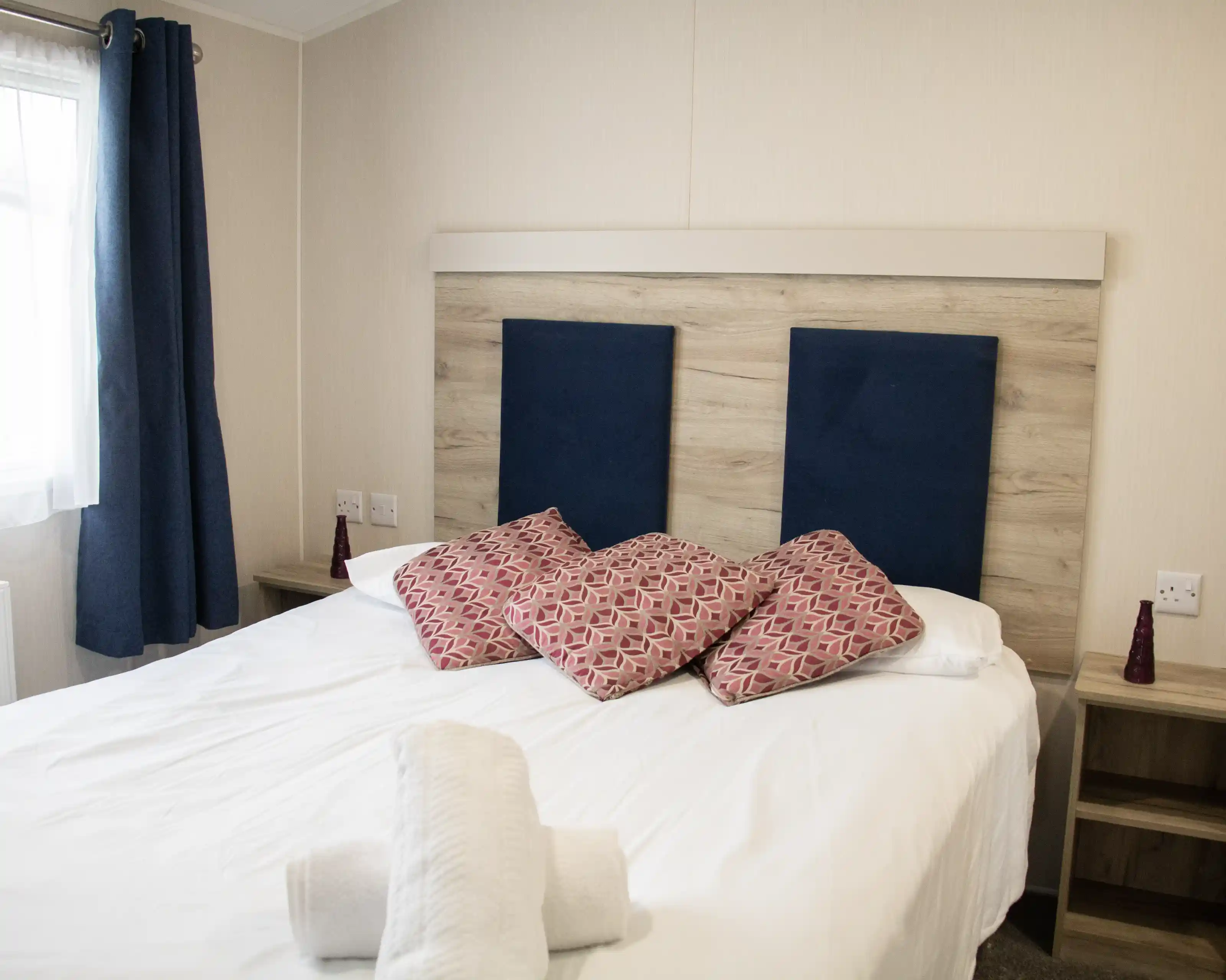 A neatly made bed with white linens, topped with decorative pillows featuring a geometric pattern. The headboard has a wood panel design with blue fabric sections. Two bedside tables with small red decorative items are visible, and curtains frame a window in the background.
