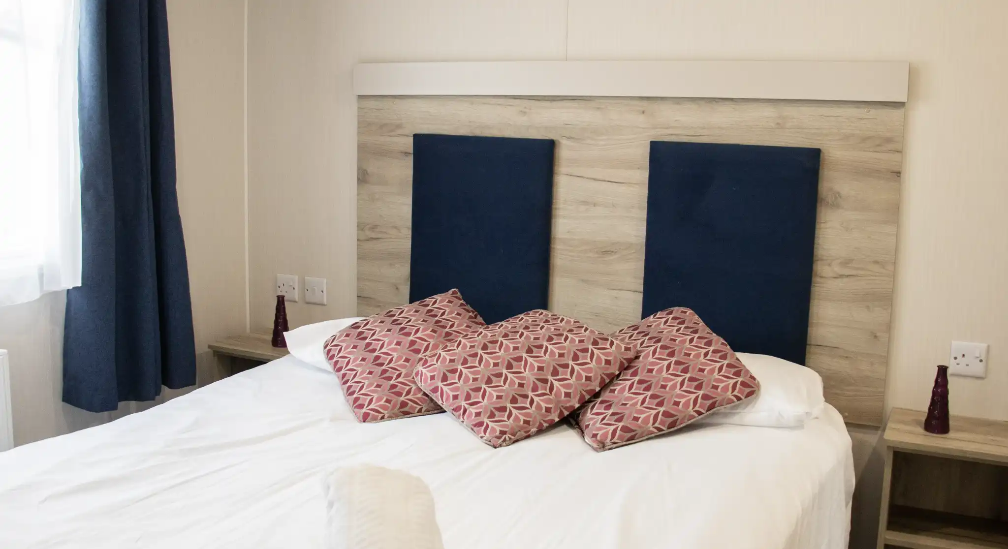 A neatly made bed with white linens, topped with decorative pillows featuring a geometric pattern. The headboard has a wood panel design with blue fabric sections. Two bedside tables with small red decorative items are visible, and curtains frame a window in the background.