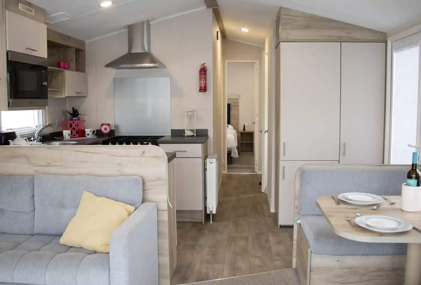 Cozy interior of a modern mobile home featuring a gray couch with a yellow pillow, a small dining area with plates, and a kitchen area with a stovetop and microwave. A bedroom door is visible in the background.