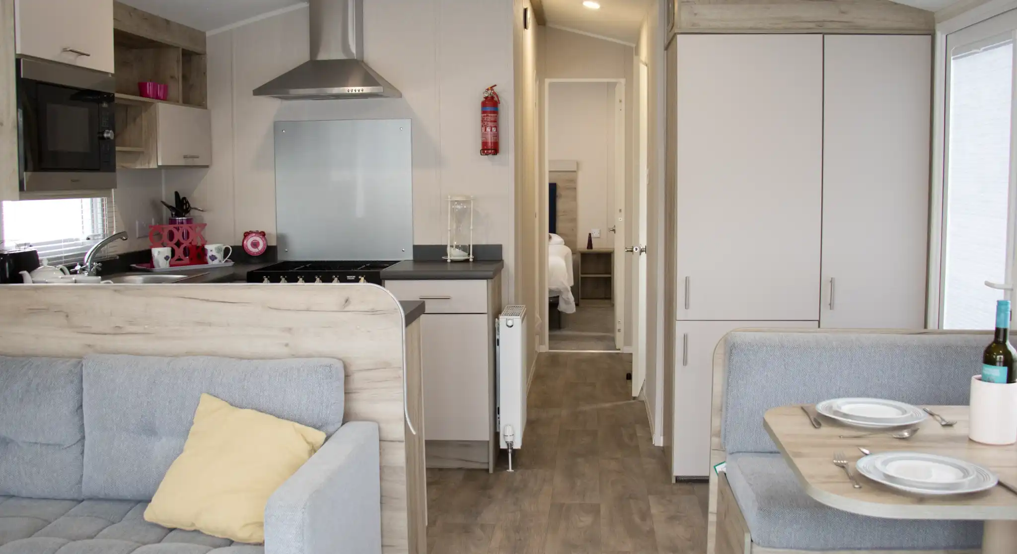 Cozy interior of a modern mobile home featuring a gray couch with a yellow pillow, a small dining area with plates, and a kitchen area with a stovetop and microwave. A bedroom door is visible in the background.