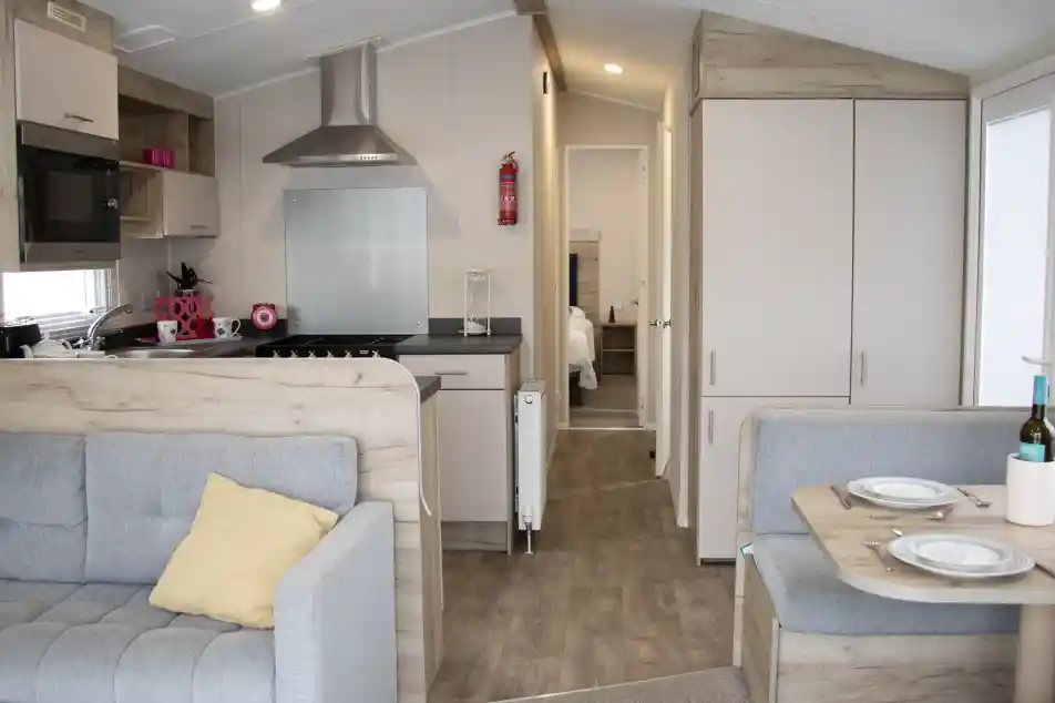 Cozy interior of a modern mobile home featuring a gray couch with a yellow pillow, a small dining area with plates, and a kitchen area with a stovetop and microwave. A bedroom door is visible in the background.