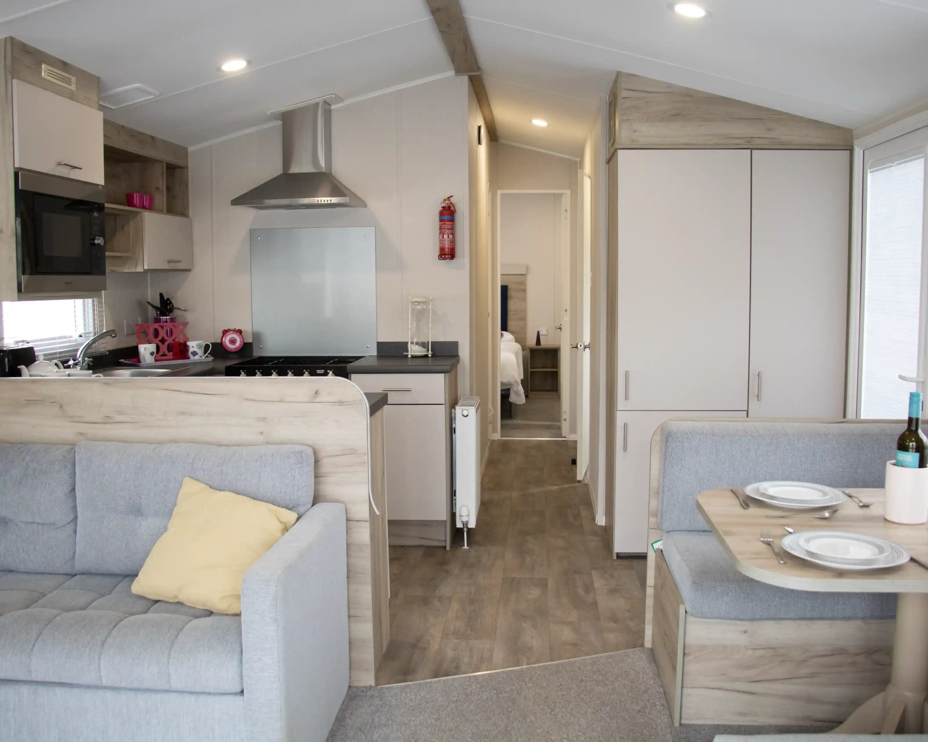 Cozy interior of a modern mobile home featuring a gray couch with a yellow pillow, a small dining area with plates, and a kitchen area with a stovetop and microwave. A bedroom door is visible in the background.