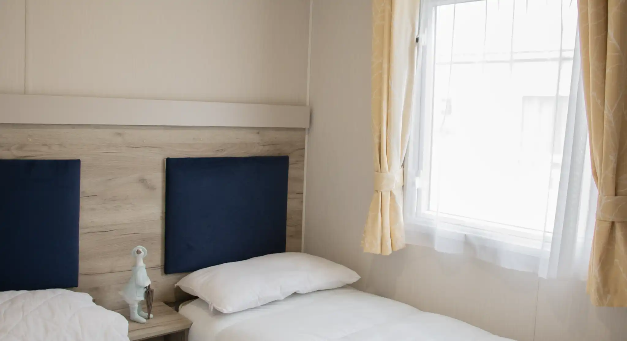 A cozy bedroom featuring two twin beds with white bedding, a wooden bedside table, and a decorative figurine. Natural light streams in through a window framed with light yellow curtains.