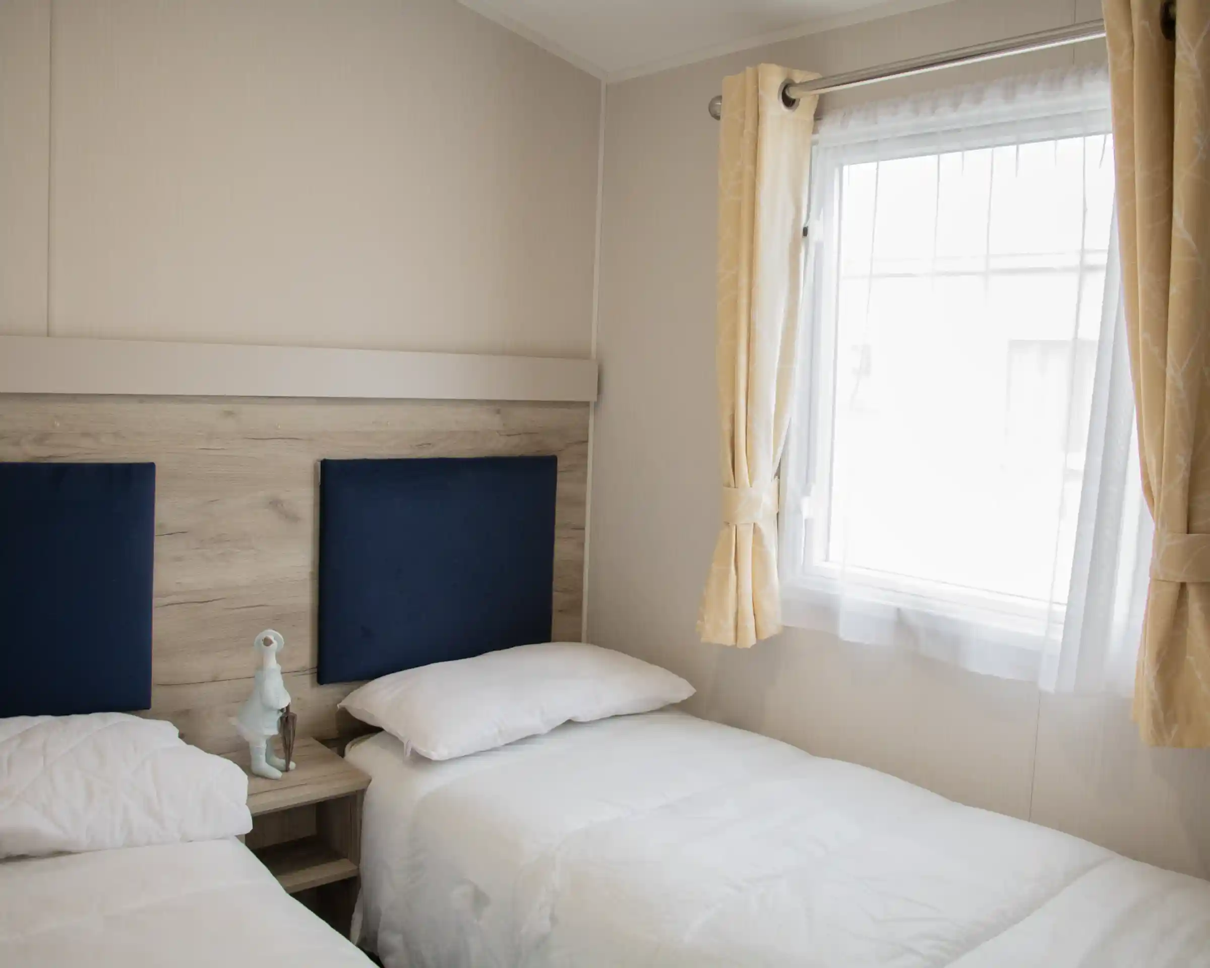 A cozy bedroom featuring two twin beds with white bedding, a wooden bedside table, and a decorative figurine. Natural light streams in through a window framed with light yellow curtains.