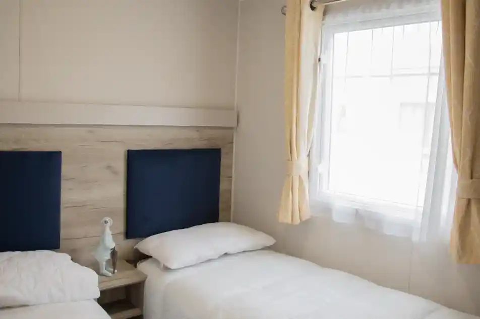 A cozy bedroom featuring two twin beds with white bedding, a wooden bedside table, and a decorative figurine. Natural light streams in through a window framed with light yellow curtains.