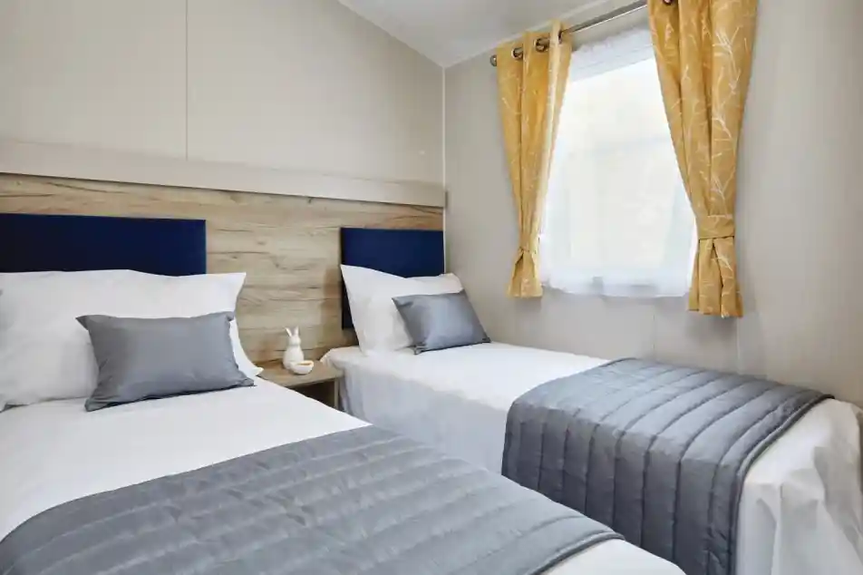 A cozy bedroom featuring two twin beds with gray bedspreads and white linens. Each bed has matching gray pillows and a wooden headboard. A small nightstand with a decorative item sits between the beds. Yellow curtains frame a window, allowing natural light to brighten the room.