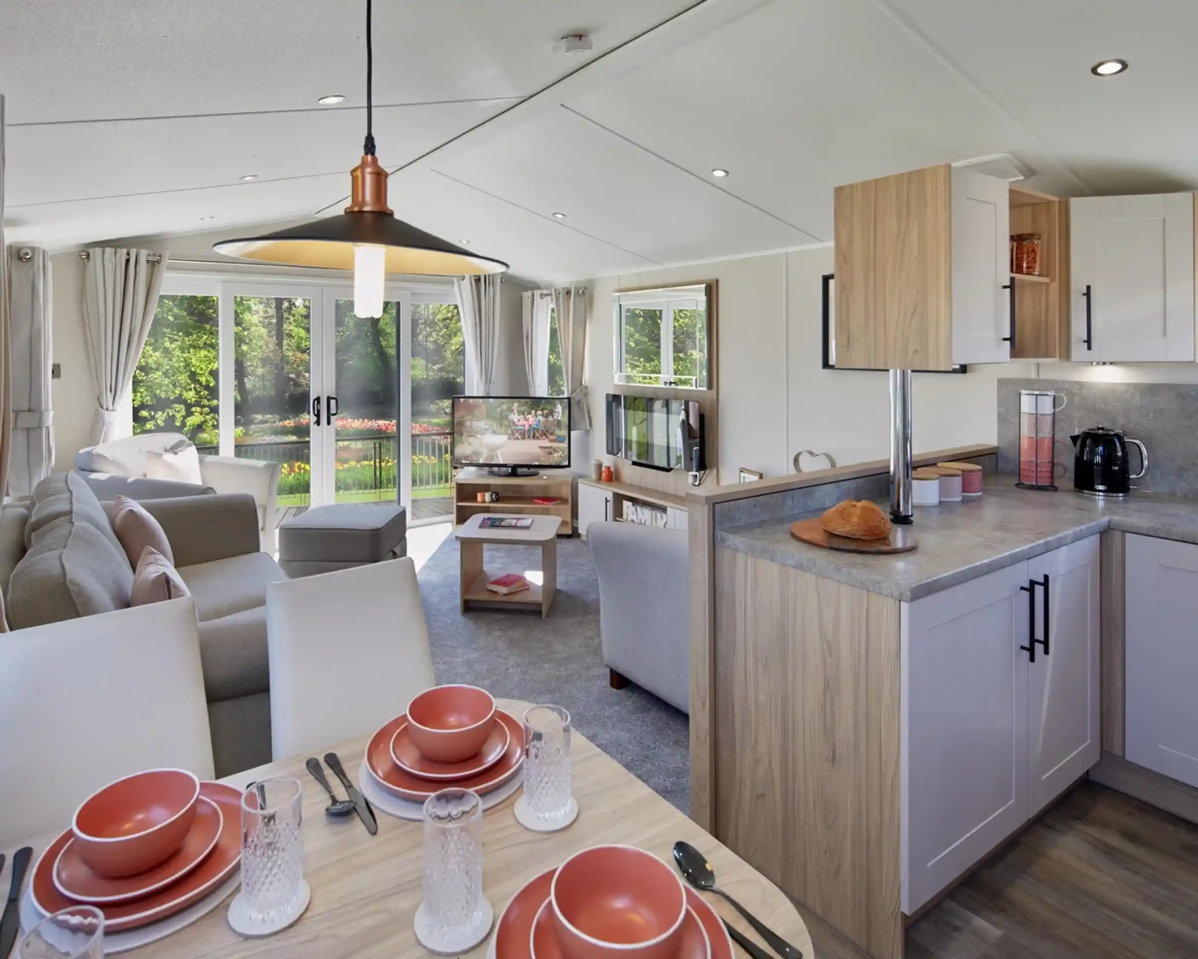 An open plan lounge, diner and kitchen with dining table set for four, kitchen cabinets and worktop, and a lounge in the background with sliding patio doors giving access outside, sofa and armchairs, a coffee table, fireplace and TV.