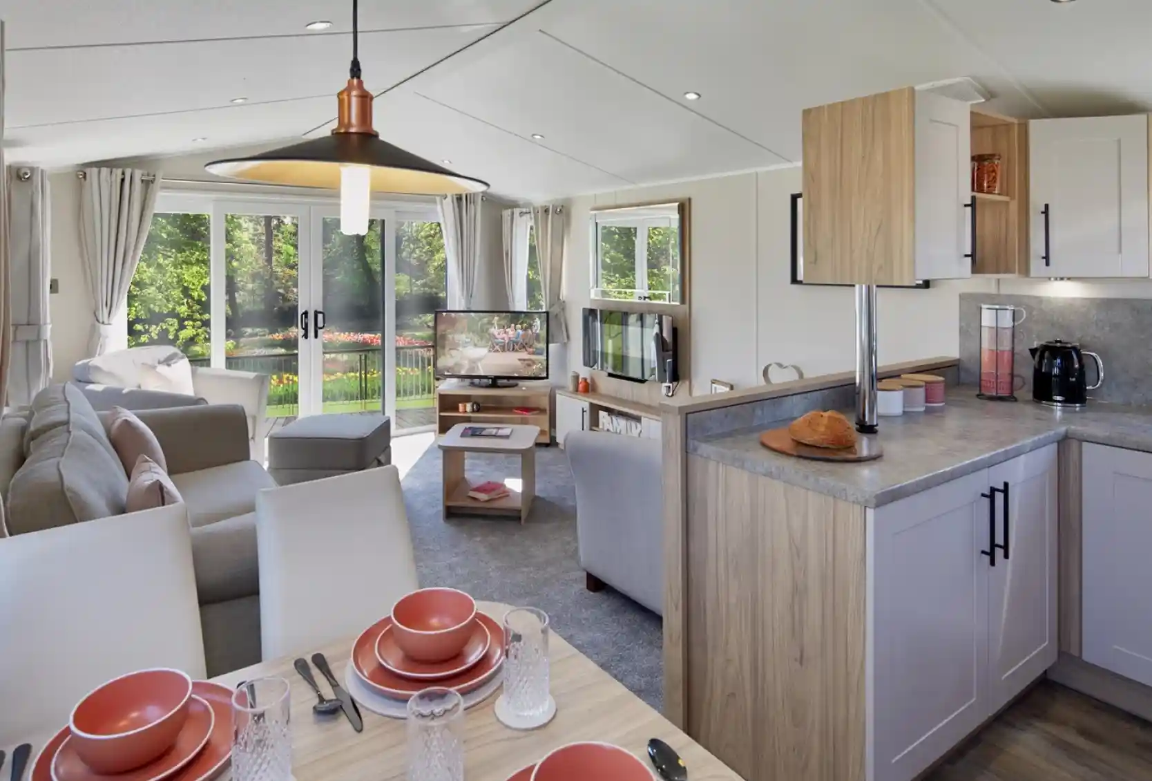 An open plan lounge, diner and kitchen with dining table set for four, kitchen cabinets and worktop, and a lounge in the background with sliding patio doors giving access outside, sofa and armchairs, a coffee table, fireplace and TV.