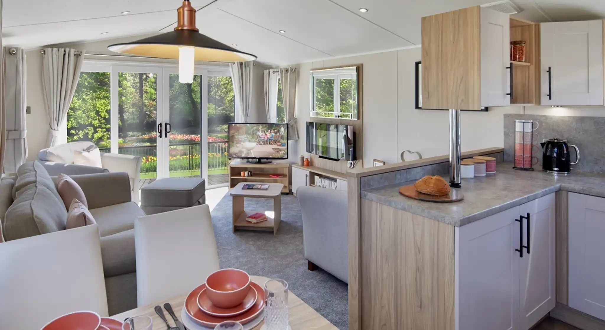 An open plan lounge, diner and kitchen with dining table set for four, kitchen cabinets and worktop, and a lounge in the background with sliding patio doors giving access outside, sofa and armchairs, a coffee table, fireplace and TV.