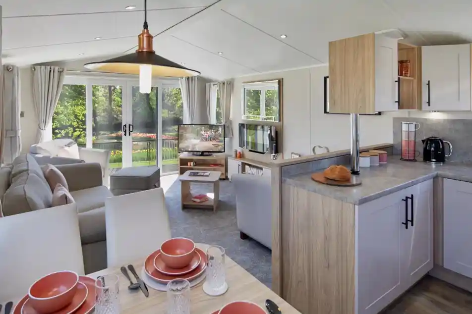 An open plan lounge, diner and kitchen with dining table set for four, kitchen cabinets and worktop, and a lounge in the background with sliding patio doors giving access outside, sofa and armchairs, a coffee table, fireplace and TV.