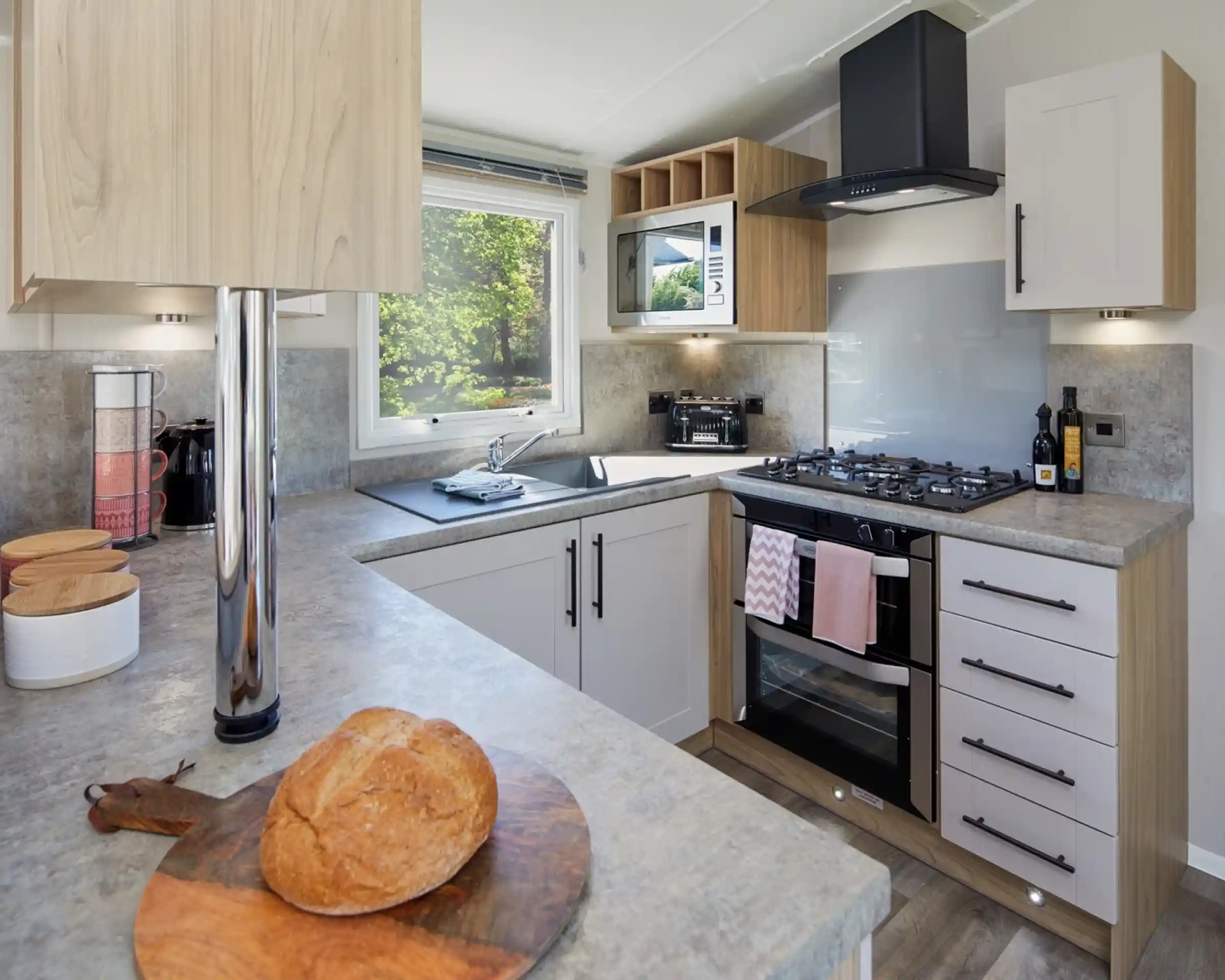 A modern U-shaped kitchen with cream and oak style cabinets and grey worktops incorporating an oven with gas hob, extractor over and resin sink unit with window over.