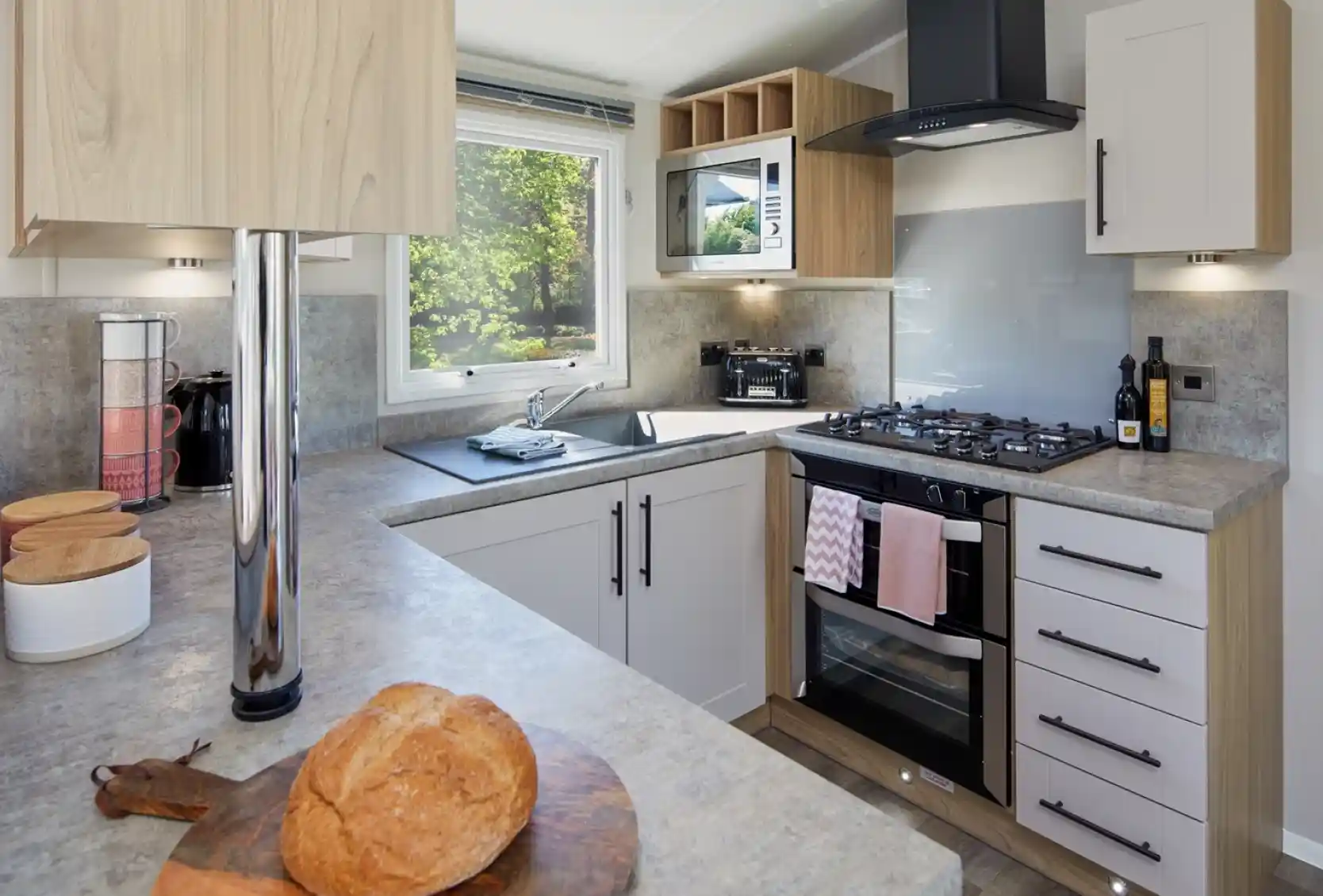 A modern U-shaped kitchen with cream and oak style cabinets and grey worktops incorporating an oven with gas hob, extractor over and resin sink unit with window over.