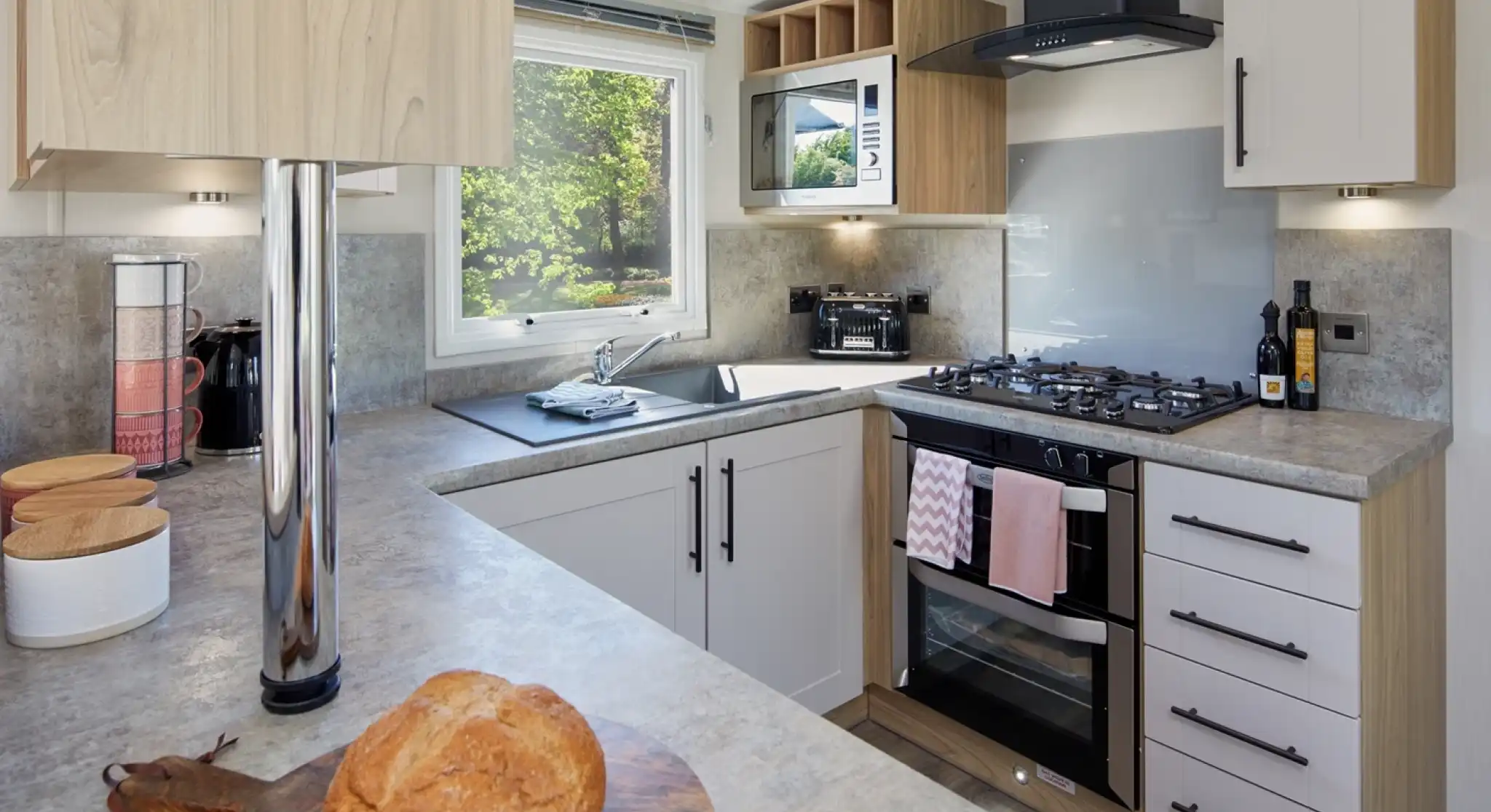 A modern U-shaped kitchen with cream and oak style cabinets and grey worktops incorporating an oven with gas hob, extractor over and resin sink unit with window over.