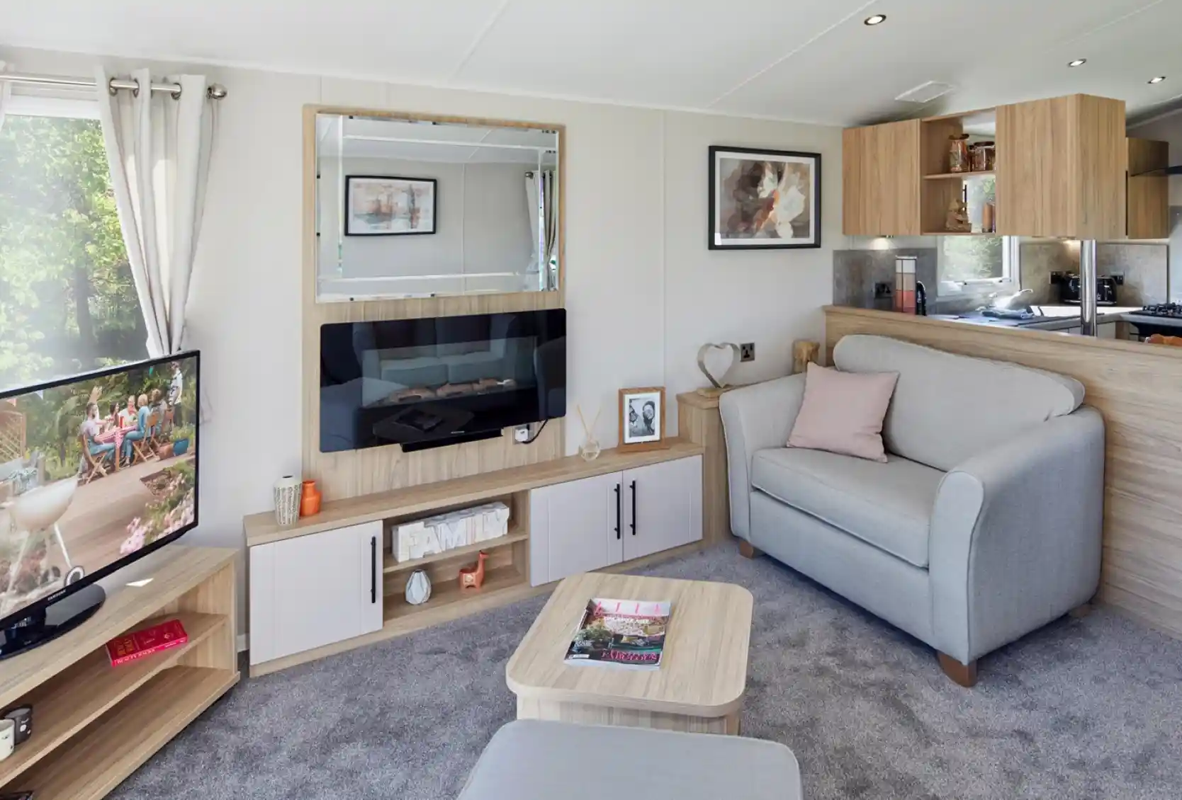 A modern lounge with feature electric fireplace with mirror above and cabinets below, together with a TV unit to one side with flat screen TV, a coffee table and a comfortable grey armchair with foot stool.