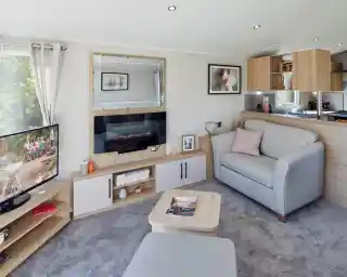 A modern lounge with feature electric fireplace with mirror above and cabinets below, together with a TV unit to one side with flat screen TV, a coffee table and a comfortable grey armchair with foot stool.