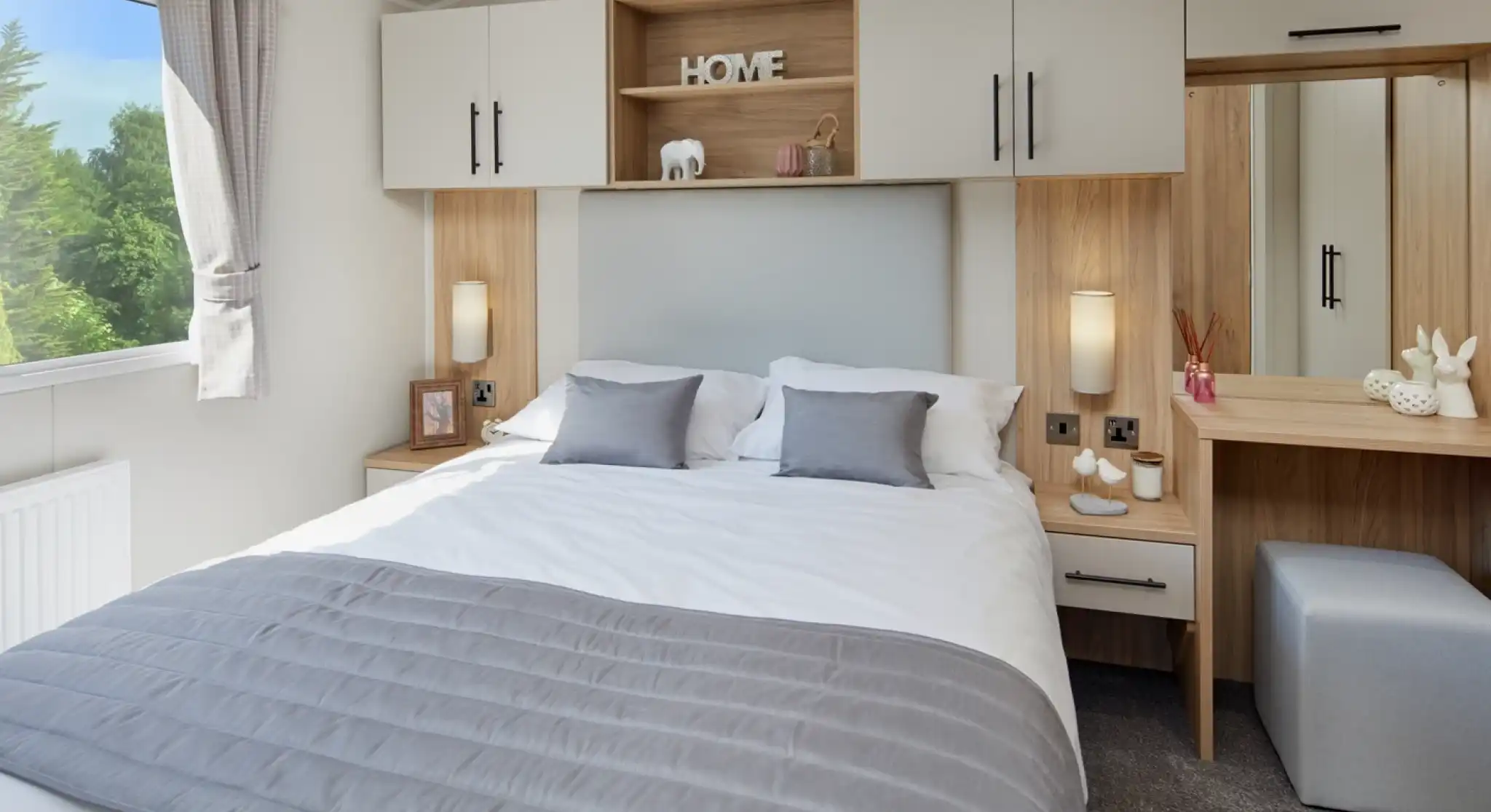 A master bedroom with double bed and built-in overhead storage, bedside units and dressing table with mirror and stool. A window gives an outlook outdoors and allows plenty of natural light.