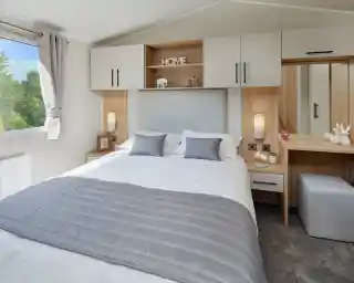 A master bedroom with double bed and built-in overhead storage, bedside units and dressing table with mirror and stool. A window gives an outlook outdoors and allows plenty of natural light.