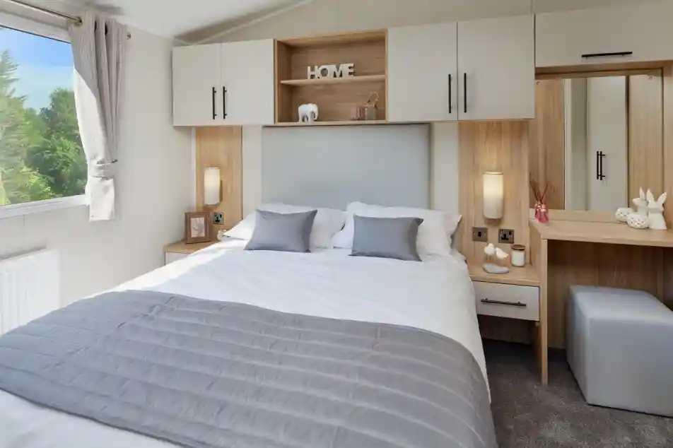 A master bedroom with double bed and built-in overhead storage, bedside units and dressing table with mirror and stool. A window gives an outlook outdoors and allows plenty of natural light.