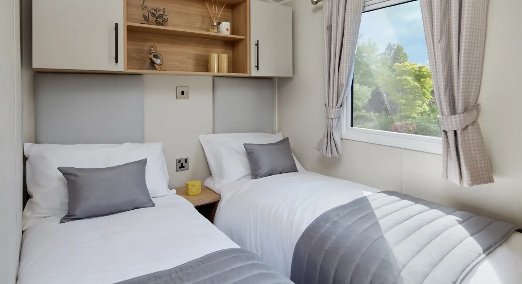 A bedroom with twin beds with crisp white sheets and pillows, together with headboards, overhead storage and a window.