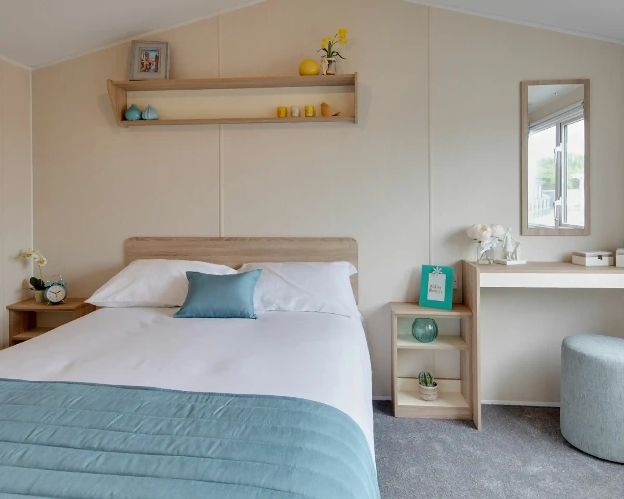 A brightly lit bedroom featuring a made bed with blue and white linens, a bedside table with an alarm clock, and a vanity with a mirror.