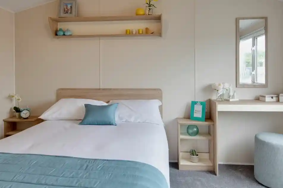 A brightly lit bedroom featuring a made bed with blue and white linens, a bedside table with an alarm clock, and a vanity with a mirror.