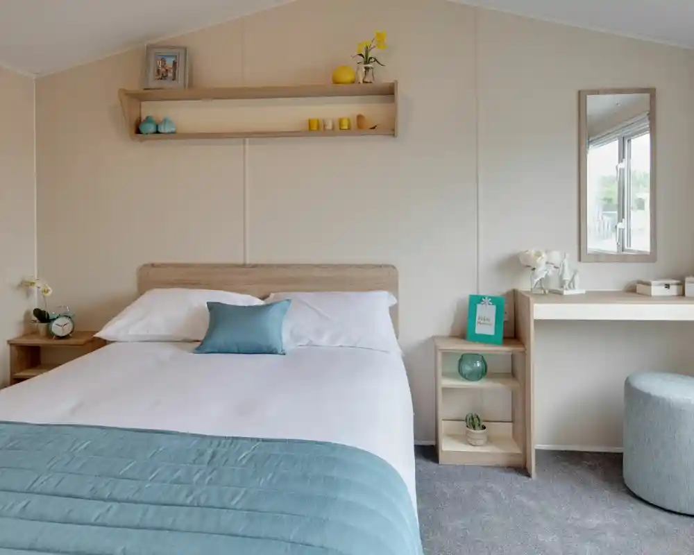A brightly lit bedroom featuring a made bed with blue and white linens, a bedside table with an alarm clock, and a vanity with a mirror.