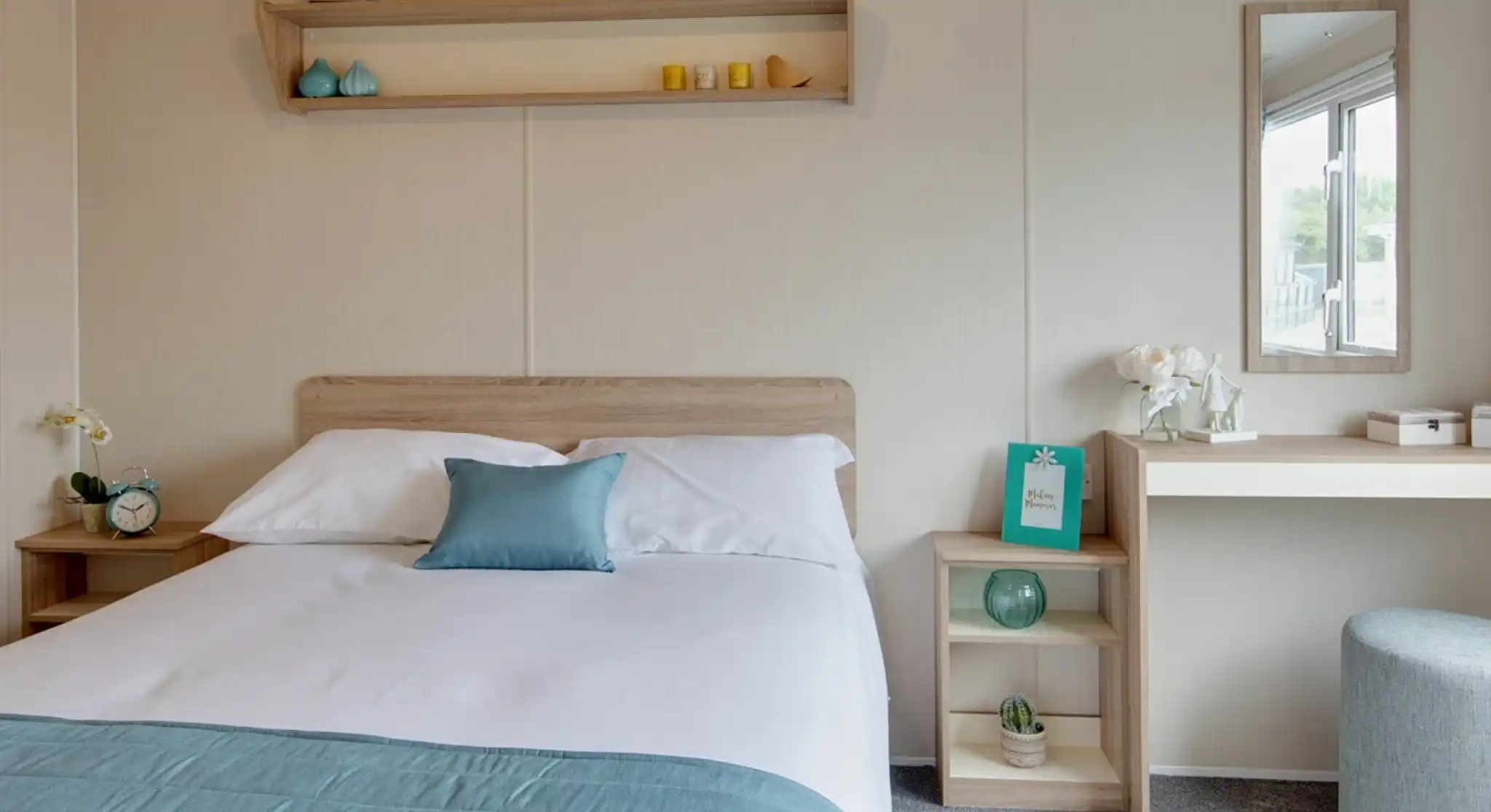A brightly lit bedroom featuring a made bed with blue and white linens, a bedside table with an alarm clock, and a vanity with a mirror.