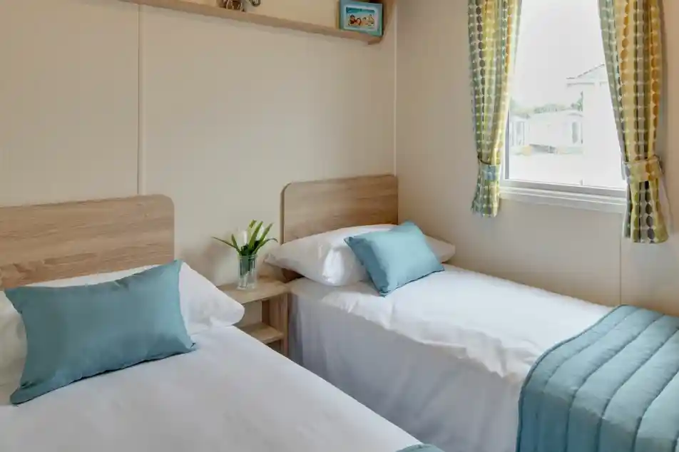 Two neatly made single beds with teal pillows and blankets sit in a room with a window and patterned curtains.