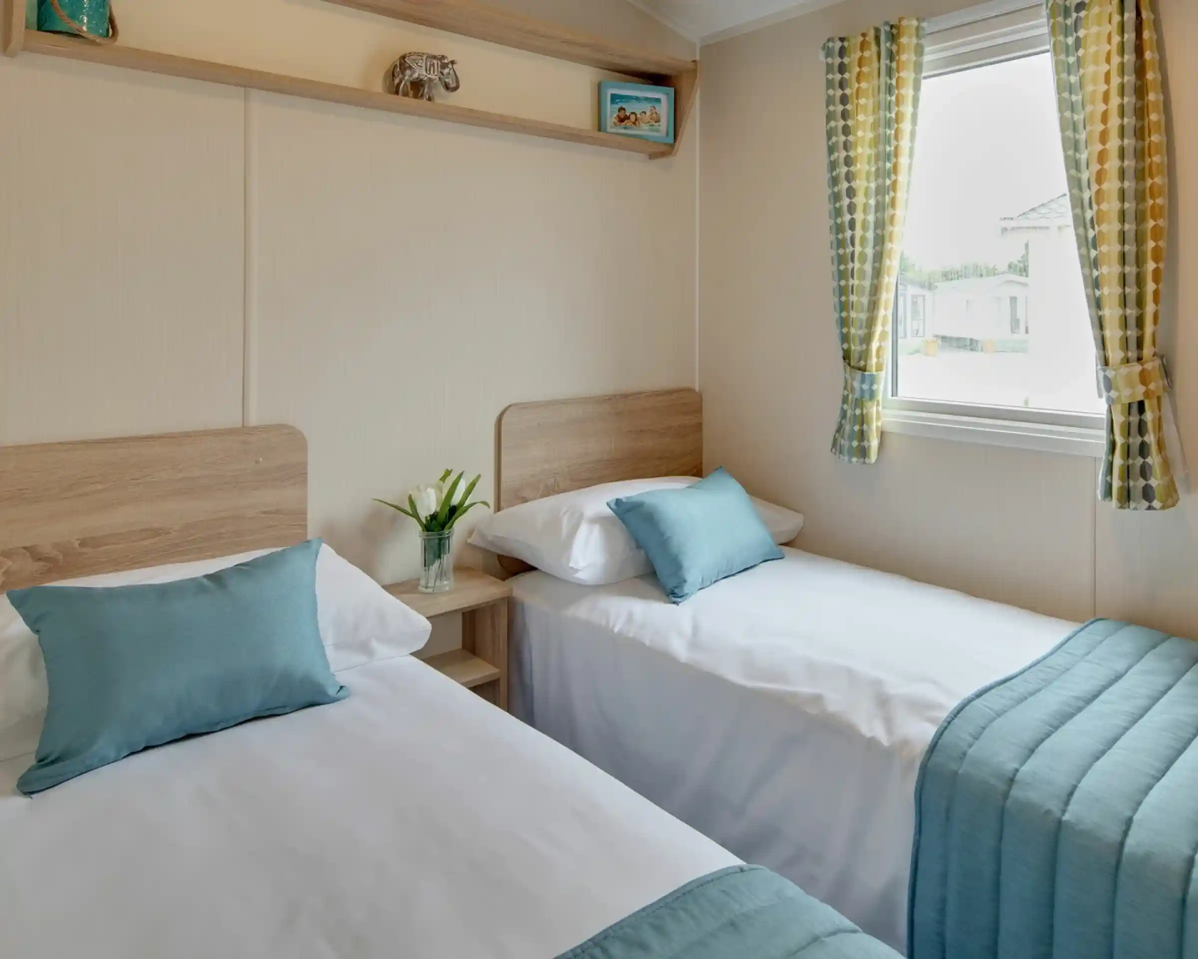 Two neatly made single beds with teal pillows and blankets sit in a room with a window and patterned curtains.