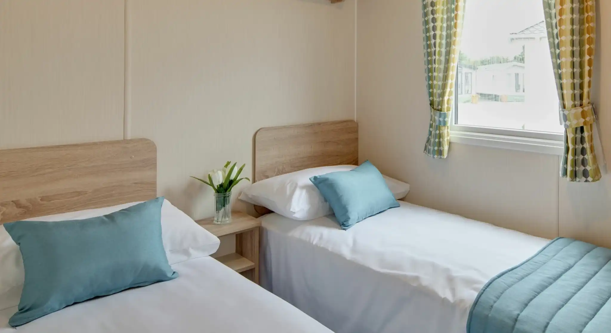 Two neatly made single beds with teal pillows and blankets sit in a room with a window and patterned curtains.