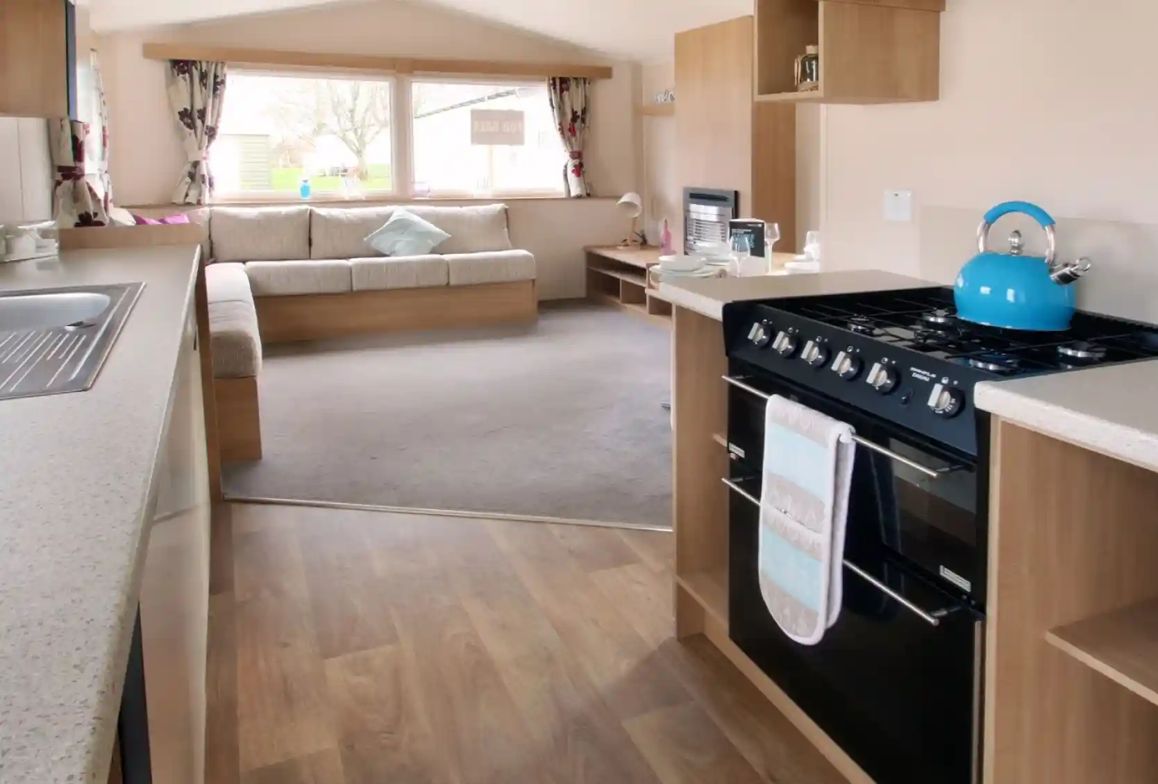 A kitchen with floor-standing over and gas hob leading into a spacious sitting room with L-shaped sofa in the background.