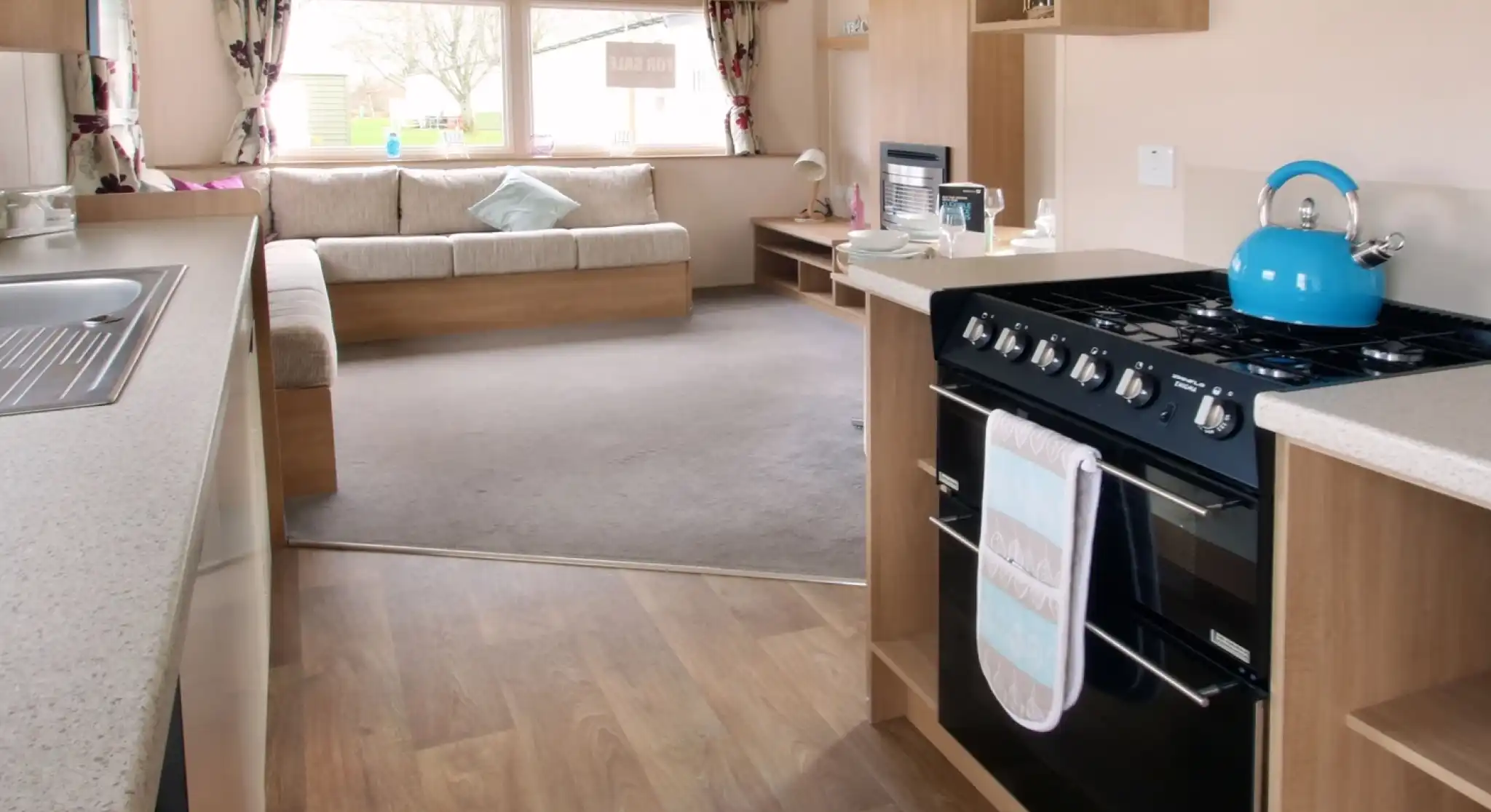 A kitchen with floor-standing over and gas hob leading into a spacious sitting room with L-shaped sofa in the background.