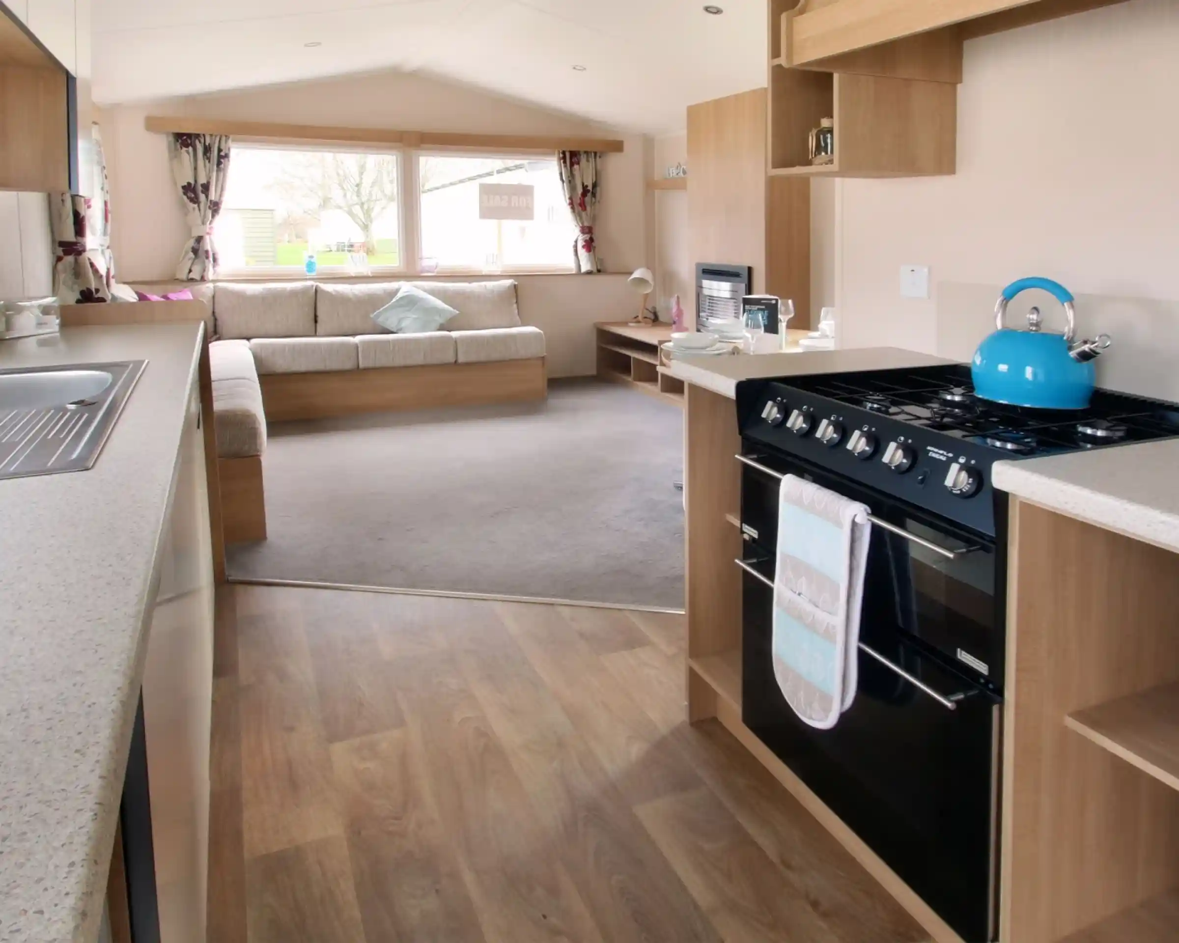 A kitchen with floor-standing over and gas hob leading into a spacious sitting room with L-shaped sofa in the background.