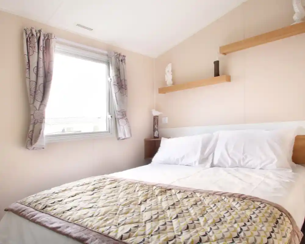 A bedroom with a double bed and patterned bed spread together with shelving and a window with floral curtains.