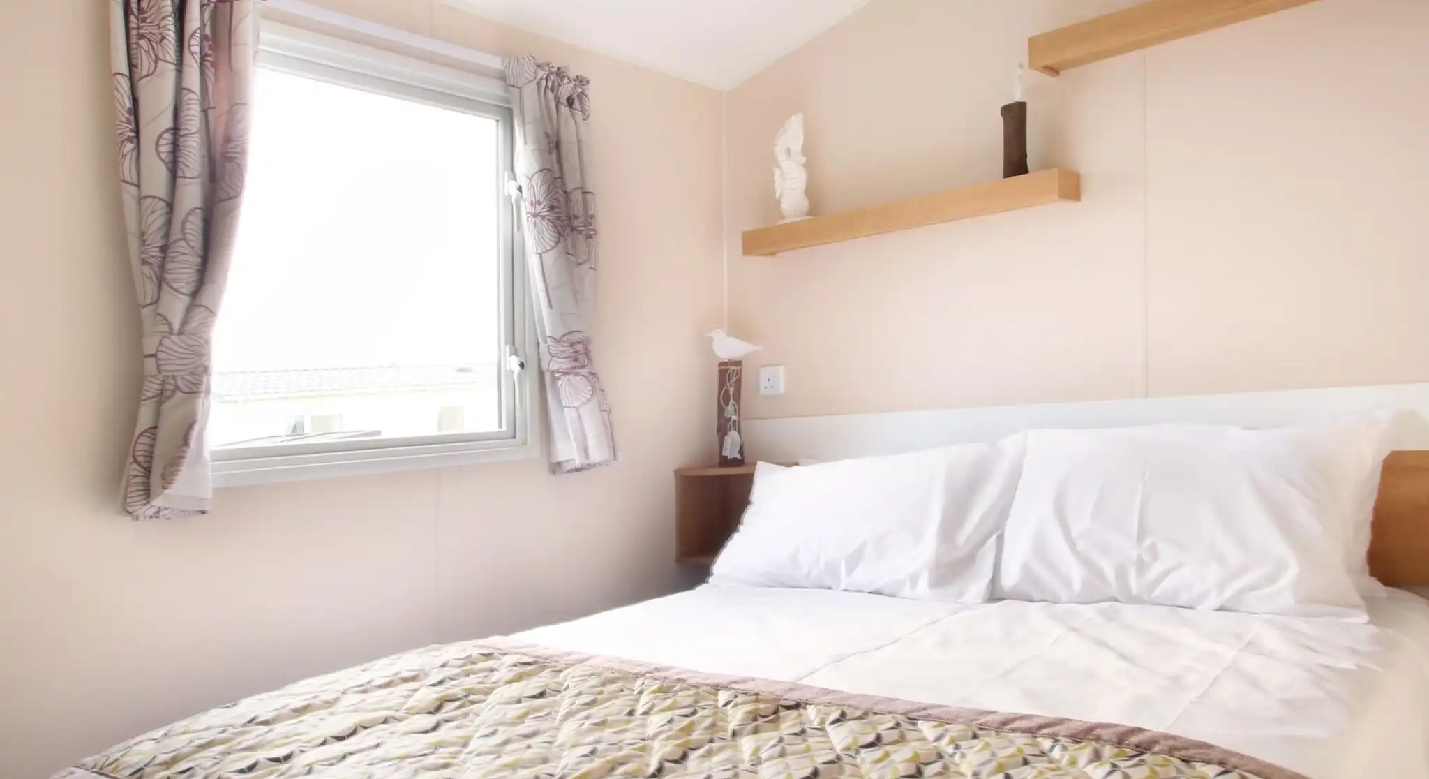 A bedroom with a double bed and patterned bed spread together with shelving and a window with floral curtains.