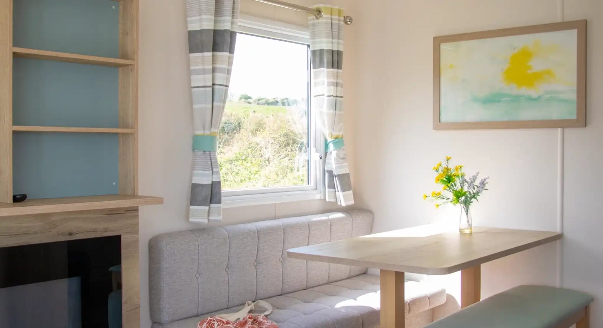 A cozy interior scene featuring a light-colored room with a gray banquette, a wooden table, and a pale turquoise ottoman. The window has sheer curtains with a striped pattern, and a bouquet of yellow flowers sits on the table. A soft, abstract painting hangs on the wall beside the window.