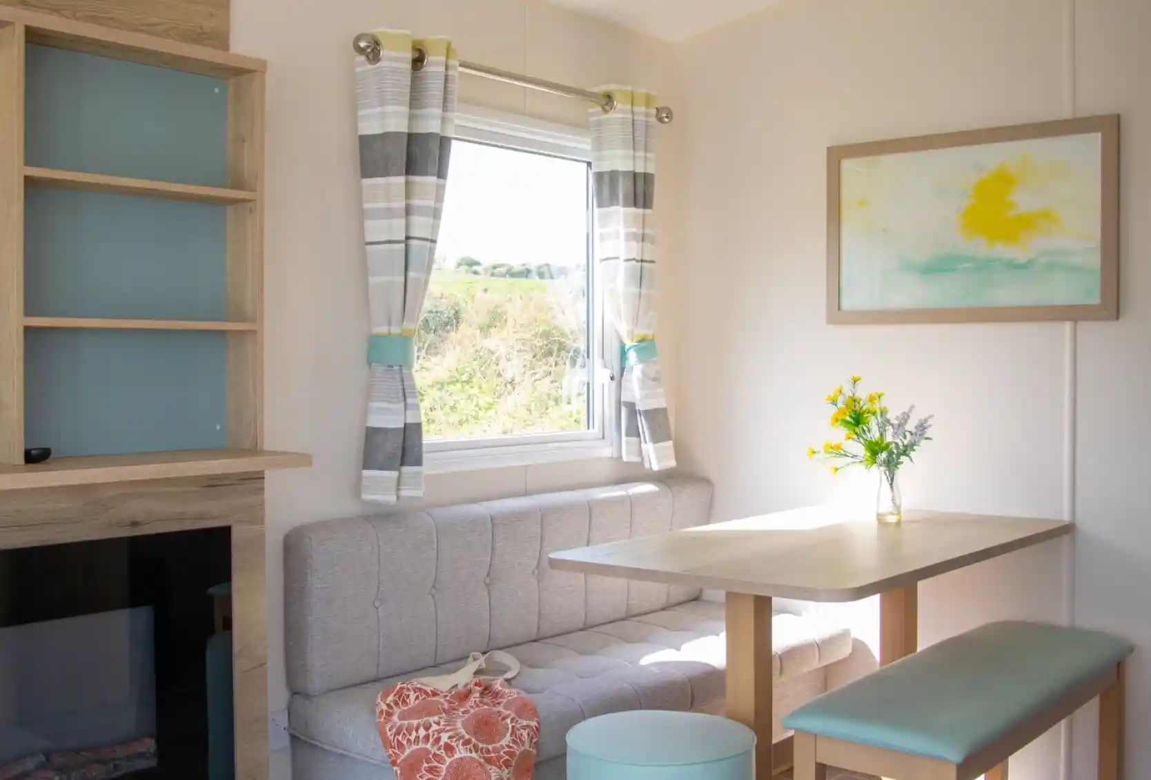 A cozy interior scene featuring a light-colored room with a gray banquette, a wooden table, and a pale turquoise ottoman. The window has sheer curtains with a striped pattern, and a bouquet of yellow flowers sits on the table. A soft, abstract painting hangs on the wall beside the window.