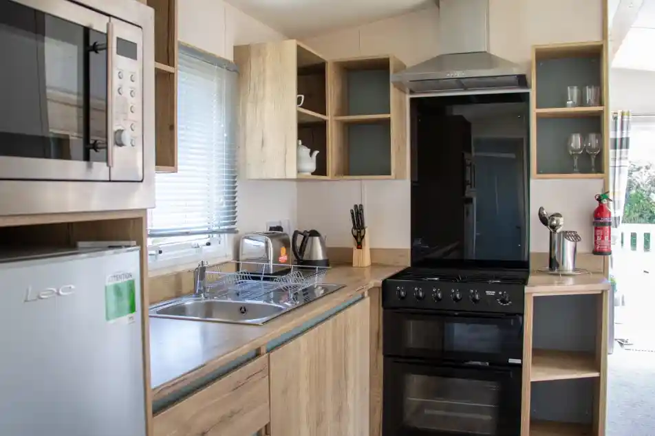 A modern kitchen featuring wooden cabinetry, a stainless steel microwave, a black oven, and a sink. A kettle and utensils are visible, along with glassware on shelves. Natural light comes through a window.