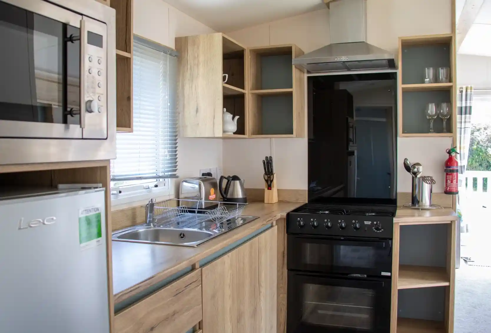 A modern kitchen featuring wooden cabinetry, a stainless steel microwave, a black oven, and a sink. A kettle and utensils are visible, along with glassware on shelves. Natural light comes through a window.
