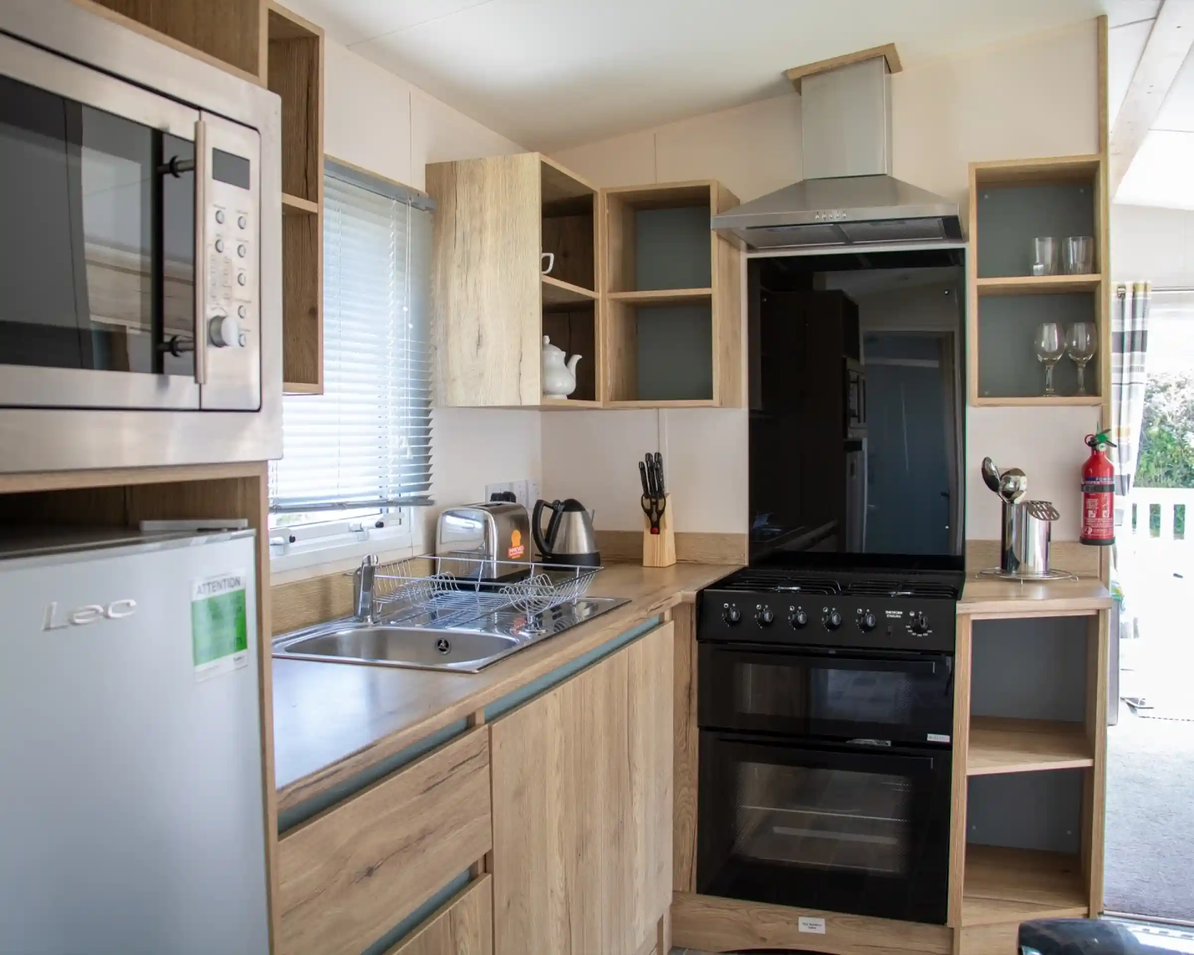A modern kitchen featuring wooden cabinetry, a stainless steel microwave, a black oven, and a sink. A kettle and utensils are visible, along with glassware on shelves. Natural light comes through a window.
