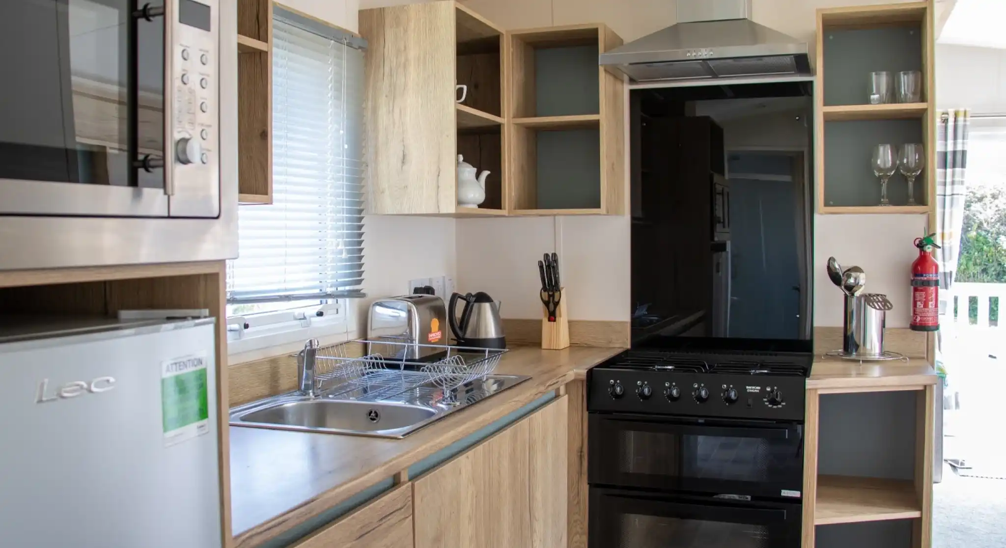 A modern kitchen featuring wooden cabinetry, a stainless steel microwave, a black oven, and a sink. A kettle and utensils are visible, along with glassware on shelves. Natural light comes through a window.