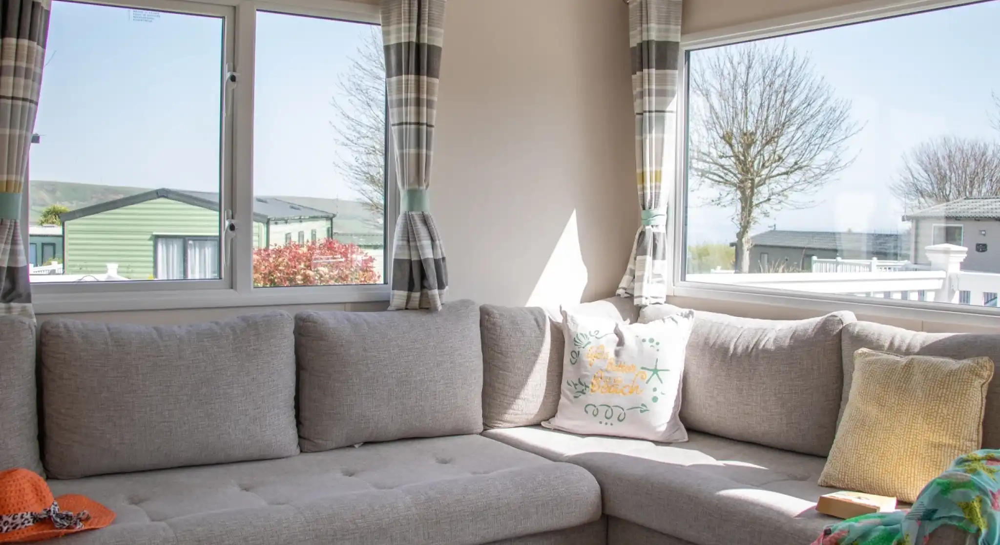 A cozy corner of a living room featuring a gray sectional sofa adorned with colorful cushions. Large windows allow natural light to brighten the space, revealing a view of nearby greenery and other mobile homes. A knitted throw and an orange hat are casually placed on the sofa.