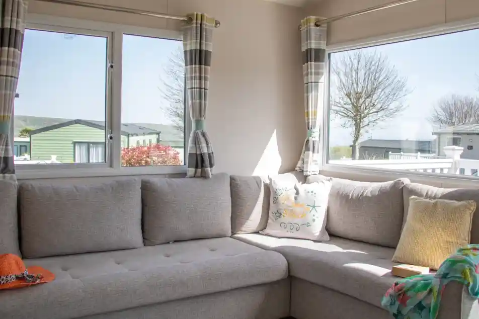 A cozy corner of a living room featuring a gray sectional sofa adorned with colorful cushions. Large windows allow natural light to brighten the space, revealing a view of nearby greenery and other mobile homes. A knitted throw and an orange hat are casually placed on the sofa.