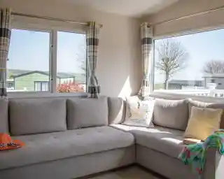 A cozy corner of a living room featuring a gray sectional sofa adorned with colorful cushions. Large windows allow natural light to brighten the space, revealing a view of nearby greenery and other mobile homes. A knitted throw and an orange hat are casually placed on the sofa.