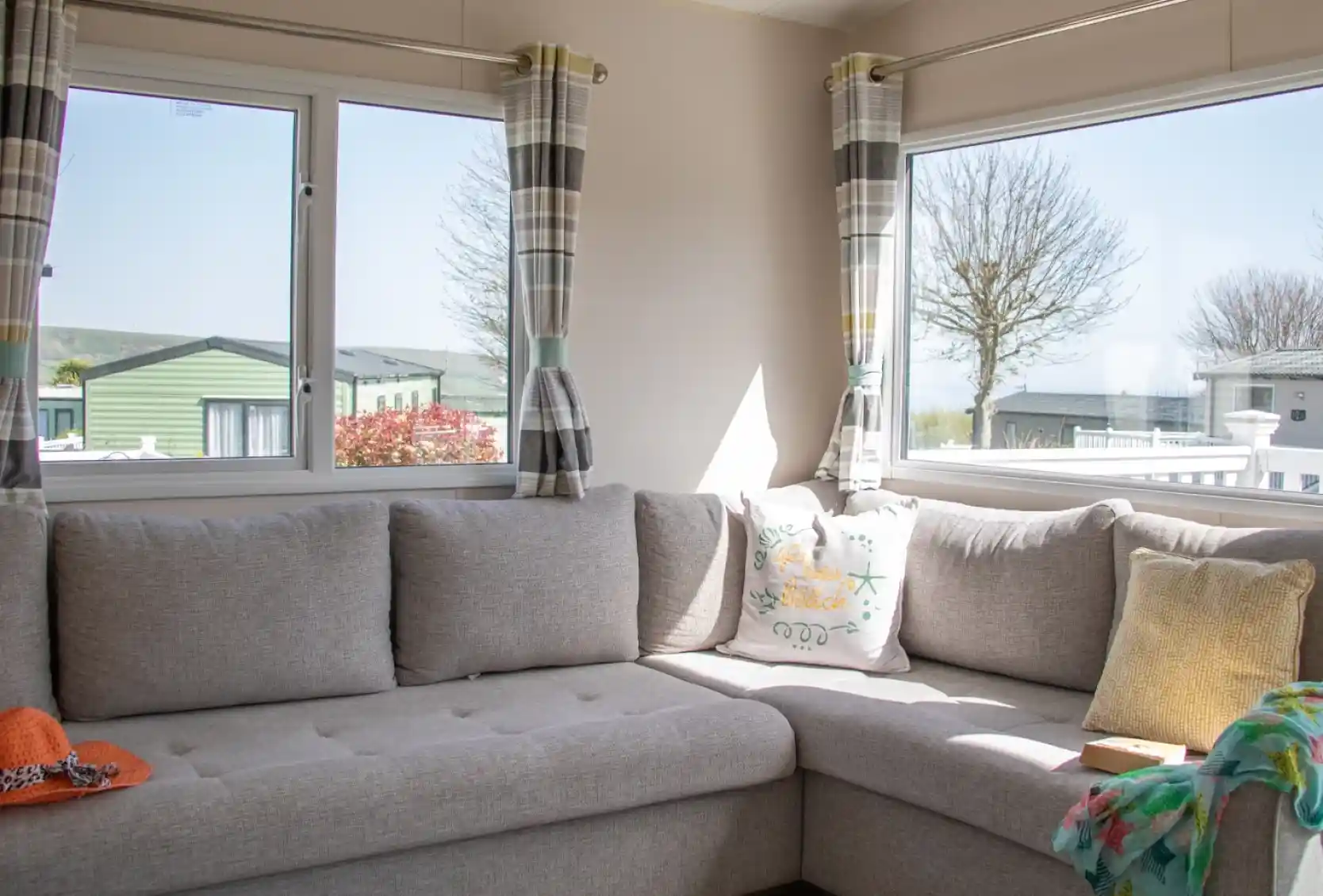 A cozy corner of a living room featuring a gray sectional sofa adorned with colorful cushions. Large windows allow natural light to brighten the space, revealing a view of nearby greenery and other mobile homes. A knitted throw and an orange hat are casually placed on the sofa.