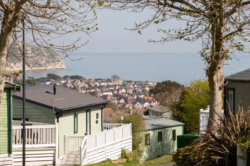 A scenic view of a coastal town from a hillside. The foreground features green and white residential buildings, with trees lining the pathway. In the background, the town's homes are visible along the water's edge, surrounded by calm sea and distant cliffs.
